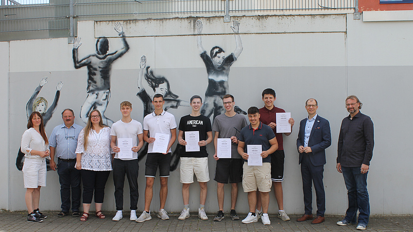 Mehrere Personen stehen vor einer Mauer mit Grafittis. Einige der Personen haben eine Urkunde in der Hand.