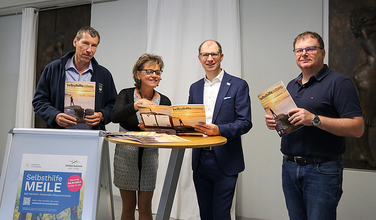 Vier Menschen bewerben die aktuelle Selbsthilfezeitung. Sie stehen an einem Tisch und ein Plakat weis auf die Selbsthilfe-MEiLE hin, die am 06.07.2024 in Bad Nauheim stattfindet.