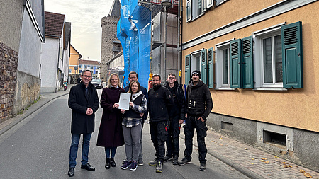 Zwei Frauen und fünf Männer stehen auf einer Straße vor einem mit einem Baugerüst eingerüsteten Gebäude. Die beiden Frauen halten gemeinsam ein Dokument in Händen.