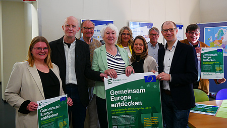 Vier Frauen und fünf Männer stehen in einem Raum. Mehrere von ihnen halten Plakate in den Händen.