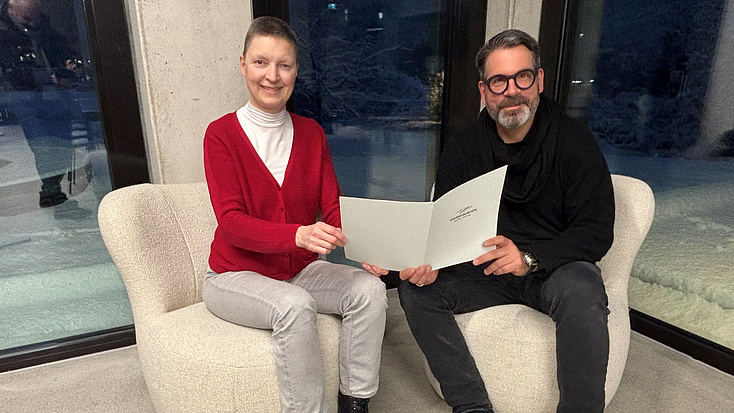 Eine Frau und ein Mann sitzen in zwei Sesseln vor einer Fensterfront. Beide halten gemeinsam ein Dokument in Händen.