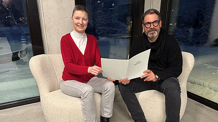 Eine Frau und ein Mann sitzen in zwei Sesseln vor einer Fensterfront. Beide halten gemeinsam ein Dokument in Händen.