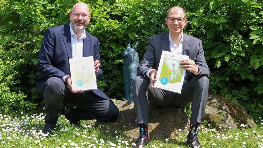 Zwei Männer sitzen vor einer grünen Hecke und halten ein Dokument in die Kamera.