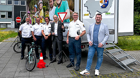 Eine Gruppe von Leuten steht vor einem Lastwagen. Um sie herum sind Verkehrsschilder, ein Fahrrad und Verkehrskegel drapiert.