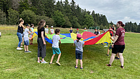 Kinder und junge Erwachsene stehen auf einer Wiese im Kreis und halten gemeinsam ein großes rundes Tuch.