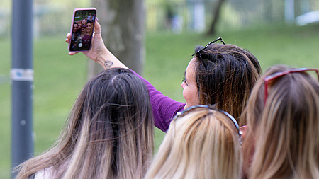 Vier junge Frauen stehen dicht gedrängt zusammen. Eine von ihnen hält ein Smartphone vor die Gruppe, um ein Selfie zu machen.