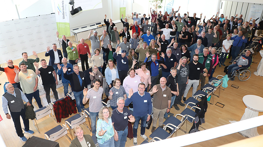 Zahlreiche Personen stehen in einem großen Saal und winken.