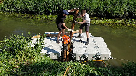 Zwei Männer stehen auf einem Floß an einem Flussufer.