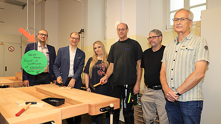 Eine Frau und fünf Männer stehen in einem Klassenraum vor einer Werkbank. Zwei Männer halten Hämmer in der Hand.