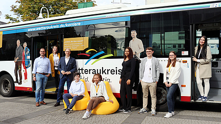Mehrere Personen stehen vor einem Bus. Auf dem Bus sind diese Personen abgebildet. Sie werben für den Wetteraukreis als Arbeitgeber.