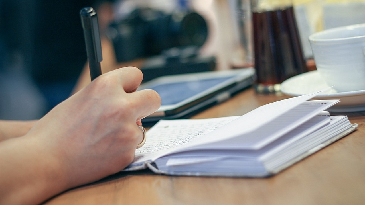 Symbolbild: Person hält Stift in der Hand, dahinter ein aufgeklapptes Buch