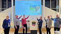 Vier Frauen und zwei Männer stehen in einem Saal und deuten auf eine Leinwand, auf der Informationen zu sehen sind.