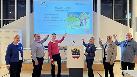 Vier Frauen und zwei Männer stehen in einem Saal und deuten auf eine Leinwand, auf der Informationen zu sehen sind.