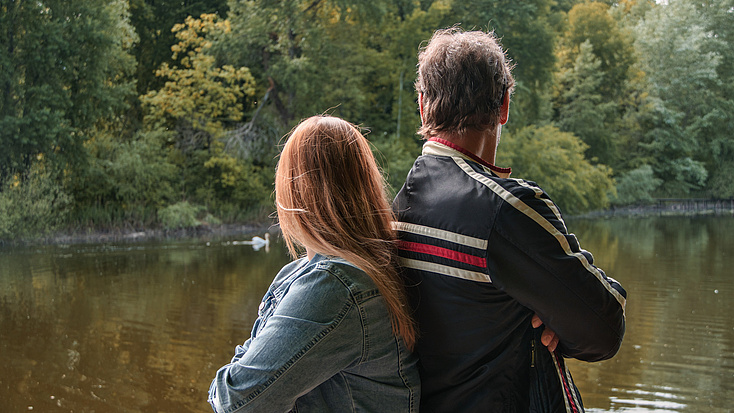 Vater und Tochter sitzen Rücken an Rücken und schauen wortlos auf einen See. Im Hintergrund sind Bäume zu sehen.