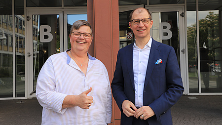 Ein Mann und eine Frau stehen lächelnd vor dem Eingang eines Gebäudes. Die Frau reckt dabei einen Daumen in die Höhe.