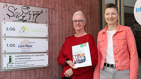 Zwei Frauen stehen vor der Eingangstür eines Gebäudes, links neben ihnen hängen mehrere Firmenschilder an der Wand. Eine der Frauen hält eine Broschüre in Händen.