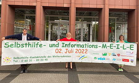 Drei Personen halten ein großes Banner,. Auf ihm steht zu lesen Selbst- und Informationsmeile, 2. Juli 2022