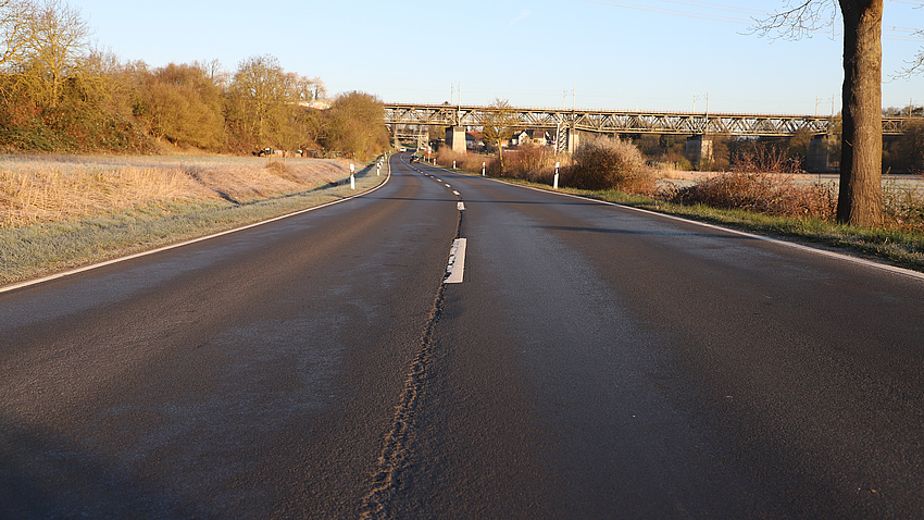 Auf der Fahrbahnoberfläche einer Straße, die über freies Feld führt, sind Straßenschäden zu erkennen. Im Bildhintergrund sieht man eine Eisenbahnbrücke, unter der die Straße hindurchführt.