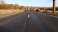 Auf der Fahrbahnoberfläche einer Straße, die über freies Feld führt, sind Straßenschäden zu erkennen. Im Bildhintergrund sieht man eine Eisenbahnbrücke, unter der die Straße hindurchführt.