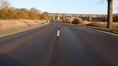 Auf der Fahrbahnoberfläche einer Straße, die über freies Feld führt, sind Straßenschäden zu erkennen. Im Bildhintergrund sieht man eine Eisenbahnbrücke, unter der die Straße hindurchführt.