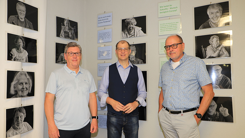 Drei Männer stehen in einem Raum vor einer Wand, an der sich mehrere Schwarz-Weiß-Fotografien und Texttafeln befinden. Auf den Fotos sind Porträts älterer Menschen zu sehen.