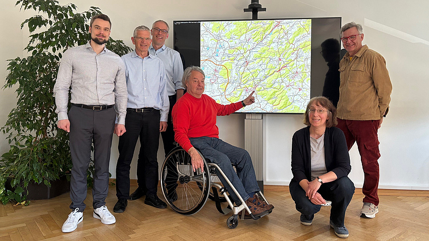 Fünf Männer, von denen einer im Rollstuhl sitzt, und eine Frau stehen in einem Raum vor einem Bildschirm. Auf diesem ist eine Landkarte zu sehen.
