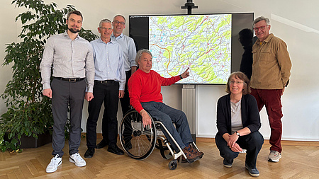 Fünf Männer, von denen einer im Rollstuhl sitzt, und eine Frau stehen in einem Raum vor einem Bildschirm. Auf diesem ist eine Landkarte zu sehen.