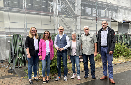 Drei Frauuen und drei Männer stehen nebeneinander auf einem Gehsteig. Im Hintergrund ein eingerüstetes Gebäude.