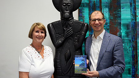 Ein Mann und eine Frau stehen neben einer großen Statue. Der Mann hält ein Buch in der Hand. An der Wand im Hintergrund ein abstraktes Gemälde