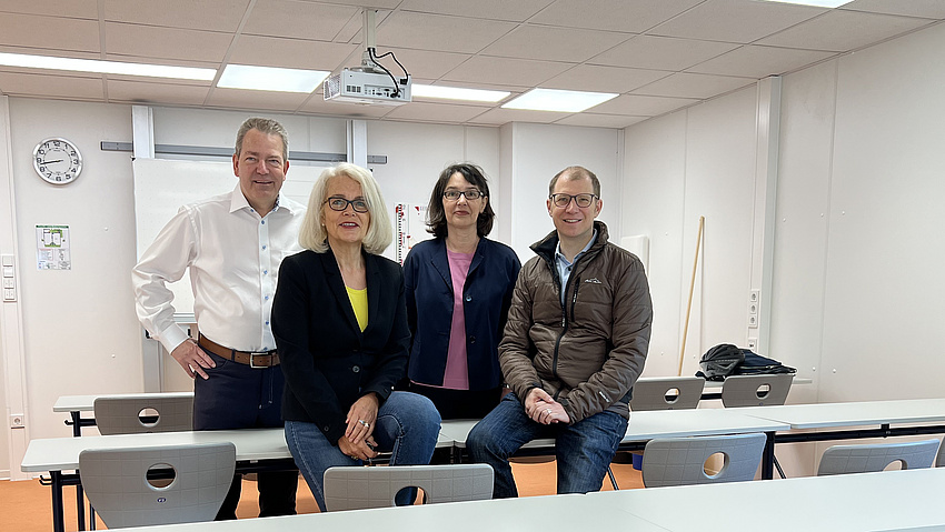 Ein Mann und eine Frau sitzen auf einer Schulbank, ein Mann und eine Frau stehen dahinter. Alle lächeln in die Kamera.
