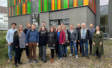 Eine Personengruppe steht vor einem Gebäude. Sie stehen nebeneinander und lächeln in die Kamera.