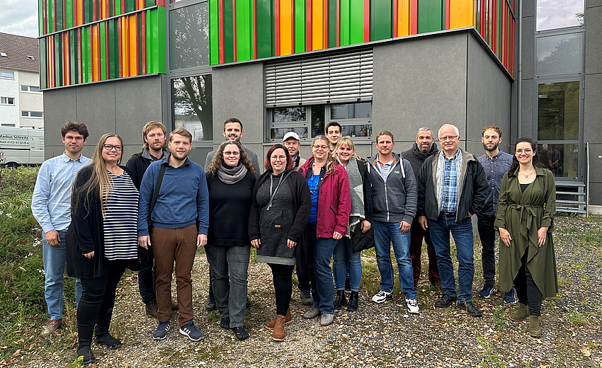 Eine Personengruppe steht vor einem Gebäude. Sie stehen nebeneinander und lächeln in die Kamera.