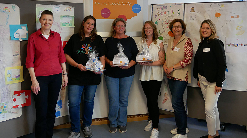 Sechs Frauen lächeln in die Kamera. Im Hintergrund gemalte Bilder an der Wand.