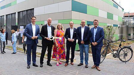 Die Personen stehen vor dem Bau mit grauen und grünen Steinen.