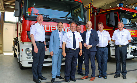 Sechs Männer stehen nebeneinander in einer Halle. Im Hintergrund zwei Löschfahrzeuge der Feuerwehr.