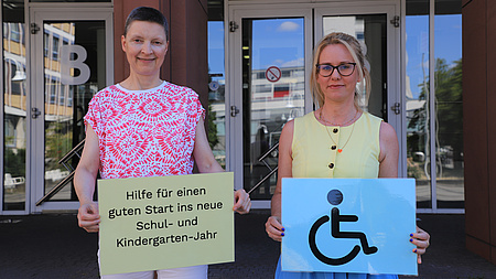 Zwei Frauen stehen vor dem Eingang eines Gebäudes und halten zwei Plakate in Händen.