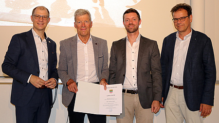 Vier Männer stehen nebeneinander. Einer von ihnen hält eine Urkunde in der Hand.