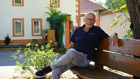 Ein Mann sitzt auf einer Holzbank unter einem Baum. Er blickt in die Kamera. Im Hintergrund ist ein altes, gepflegtes Gebäude zu erkennen.