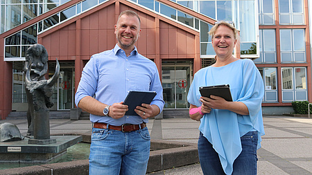 Ein Mann und eine Frau stehen nebeneinander vor einem Gebäude. Sie halten ein Notepad in der Hand. Im Hintergrund ein Brunnen mit einer weiblichen Figur.