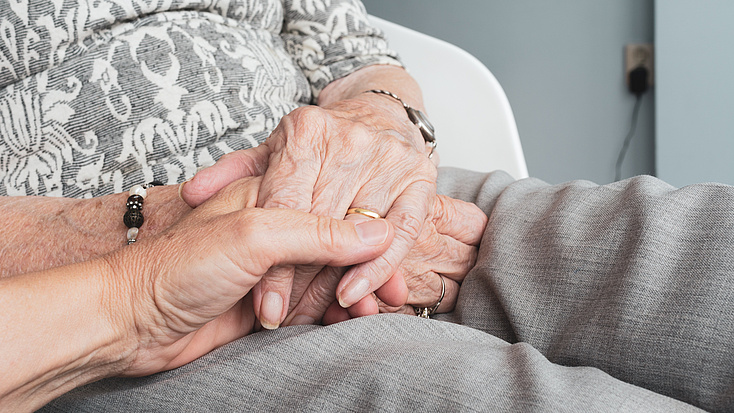 Eine alte Frau sitzt in einem Sessel, während eine jüngere Person ihre Hand hält.