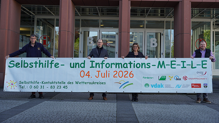 Eine Frau und drei Männer stehen vor dem Eingang eines großen Gebäudes und halten gemeinsam ein großes Transparent in Händen.