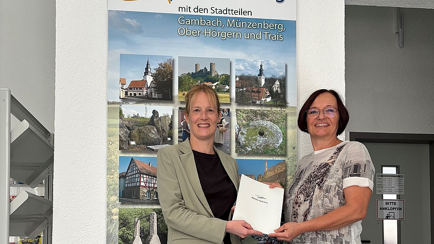 Zwei Frauen stehen in einem Raum vor einem Plakat, das verschiedene Ortsansichten zeigt. Beide halten gemeinsam ein Dokument in Händen.