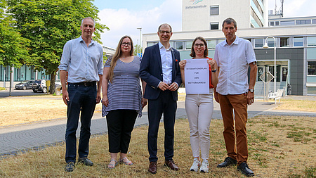 Zwei Frauen und drei Männer stehen auf einer trockenen Wiese. Eine der beiden Frauen hält ein Papier in die Kamera. Auf diesem steht: Hitzeaktionsplan für den Wetteraukreis