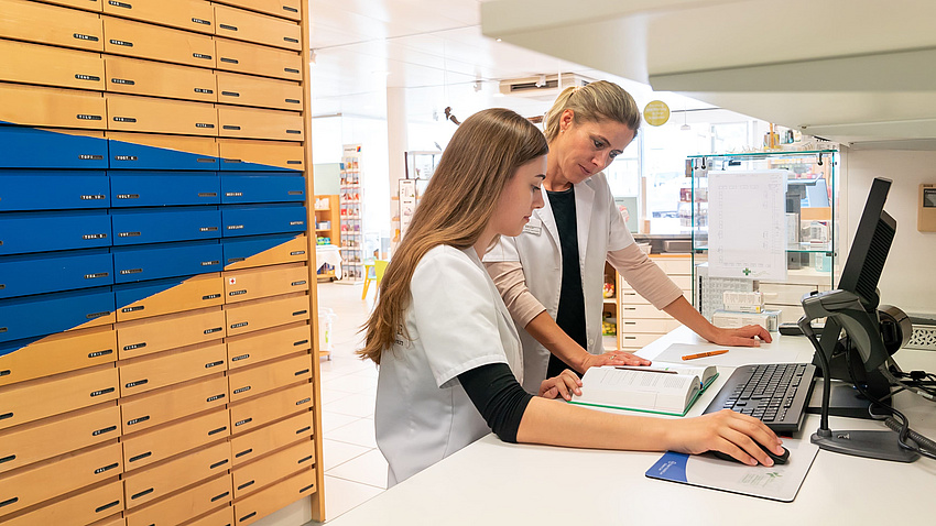 Eine Frau und eine Jugendliche stehen in einem Apothekenraum gemeinsam an einem Tisch. Beide tragen weiße Kittel. Vor ihnen liegt ein aufgeschlagenes Buch und daneben steht ein PC, in dem beide etwas nachsehen.