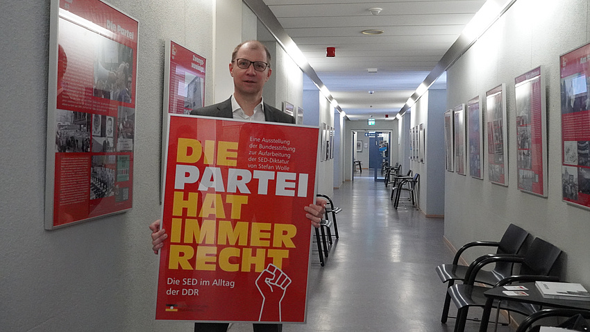 Ein Mensch steht vor einer langen Wand, er hält ein Plakat in den Händen.