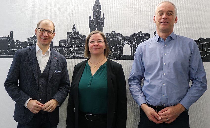Zwei Männer und eine Frau stehen nebeneinander und lächeln in die Kamera. An der Wand im Hintergrund ein Scherenschnitt mit Häusern und Türmen.