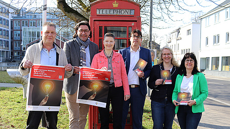Mehrere Personen stehen vor einer Telefonzelle und halten Plakate und Flyer in den Händen mit der Aufschrift Bildungsmesse Wetterau.