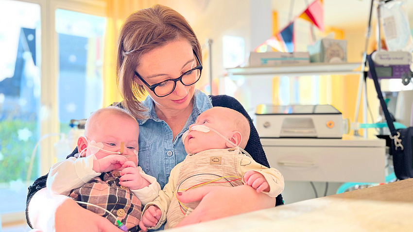 Eine junge Frau hält zwei beatmete Säuglinge auf dem Arm.
