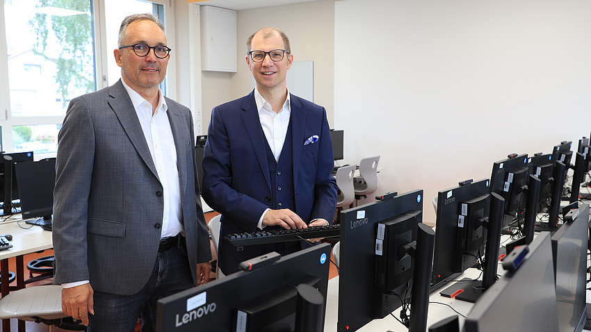 Zwei Männer stehen in einem Raum vor einer Anzahl Computer-Bildschirme. Einer der Männer hält zudem eine Tastatur in den Händen.