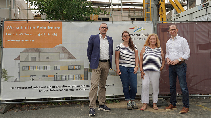 Vier Menschen stehen vor einem Bauzaun, im Hintergrund ist eine Großbaustelle.